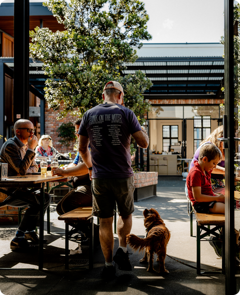 Man with a dog walking past people at outdoor tables in a casual setting at Brave Brewing Co
