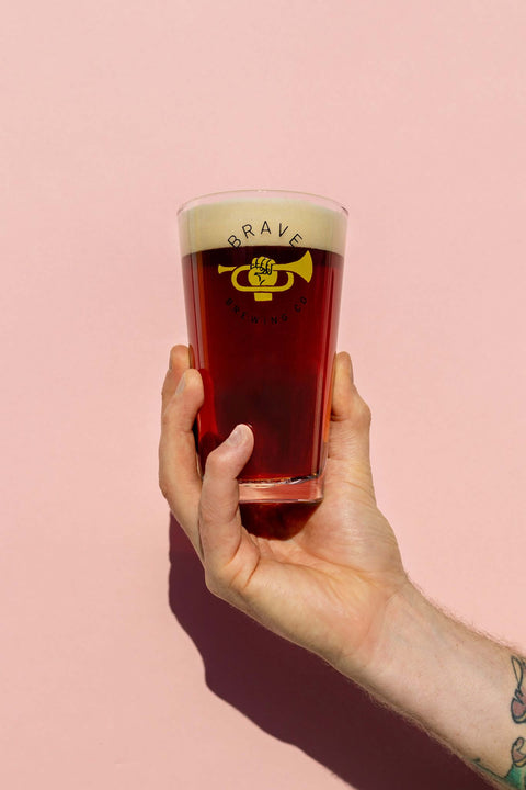Hand holding a glass of beer with 'Brave Brewing Co' branding against a pink background