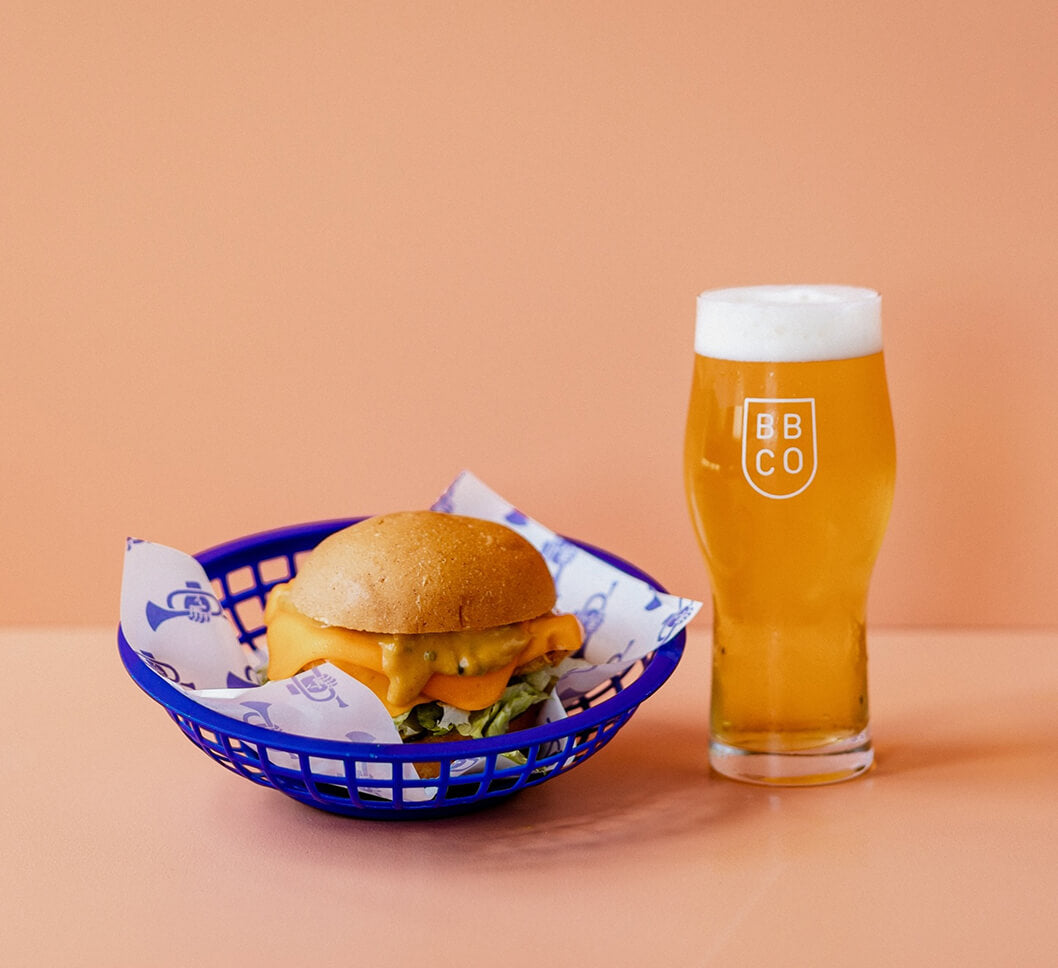 Burger in a Brave brewing Co blue basket with a glass of beer on a peach background