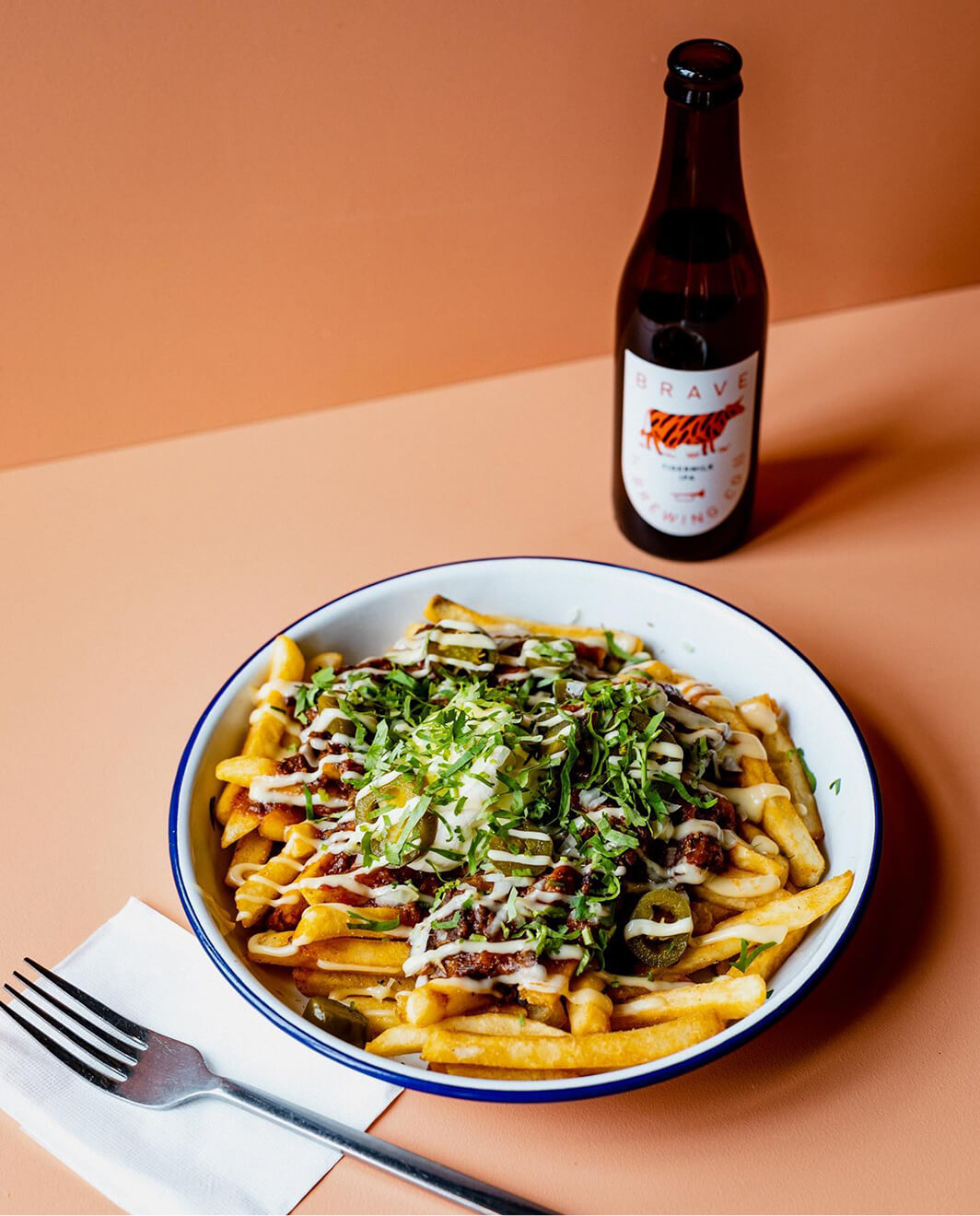 Bowl of Poutine fries with toppings and a bottle of beer on a peach-colored background