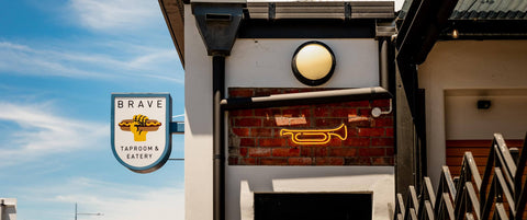 Brave brewing Co Taproom & Eatery exterior with neon sign and logo against a blue sky.