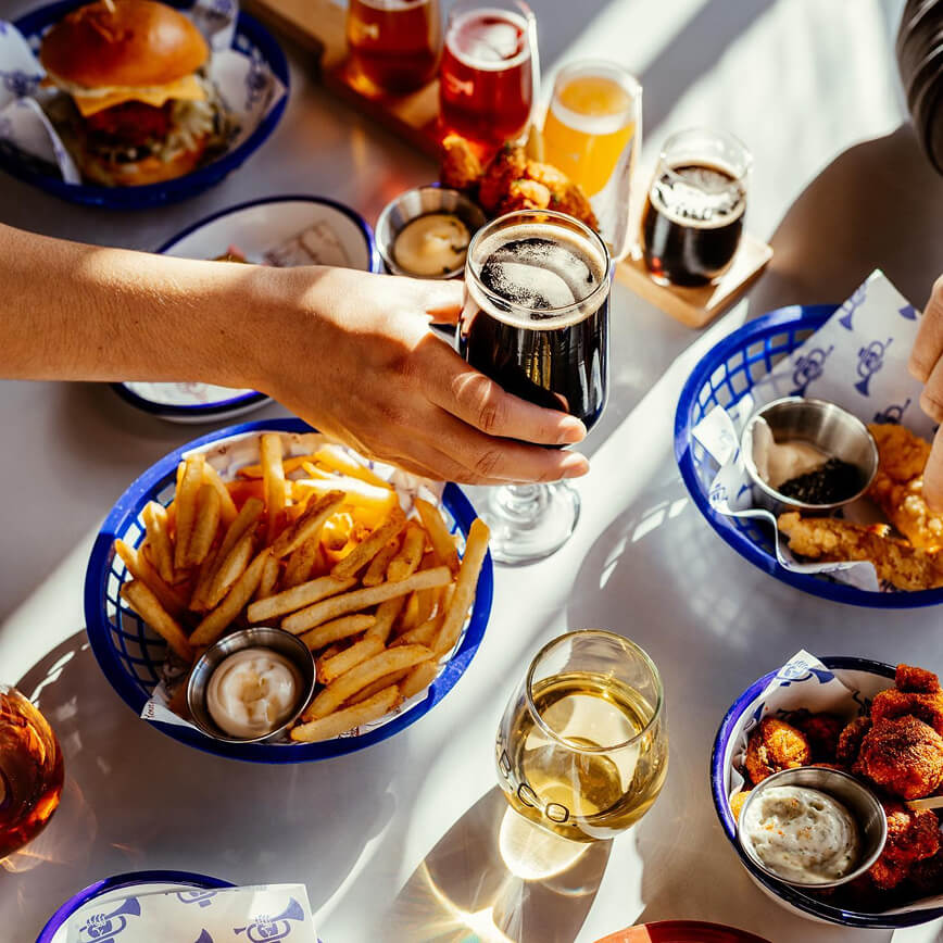 Table with food and drinks including burgers, fries, and glasses of beer and wine at Brave Brewing Co