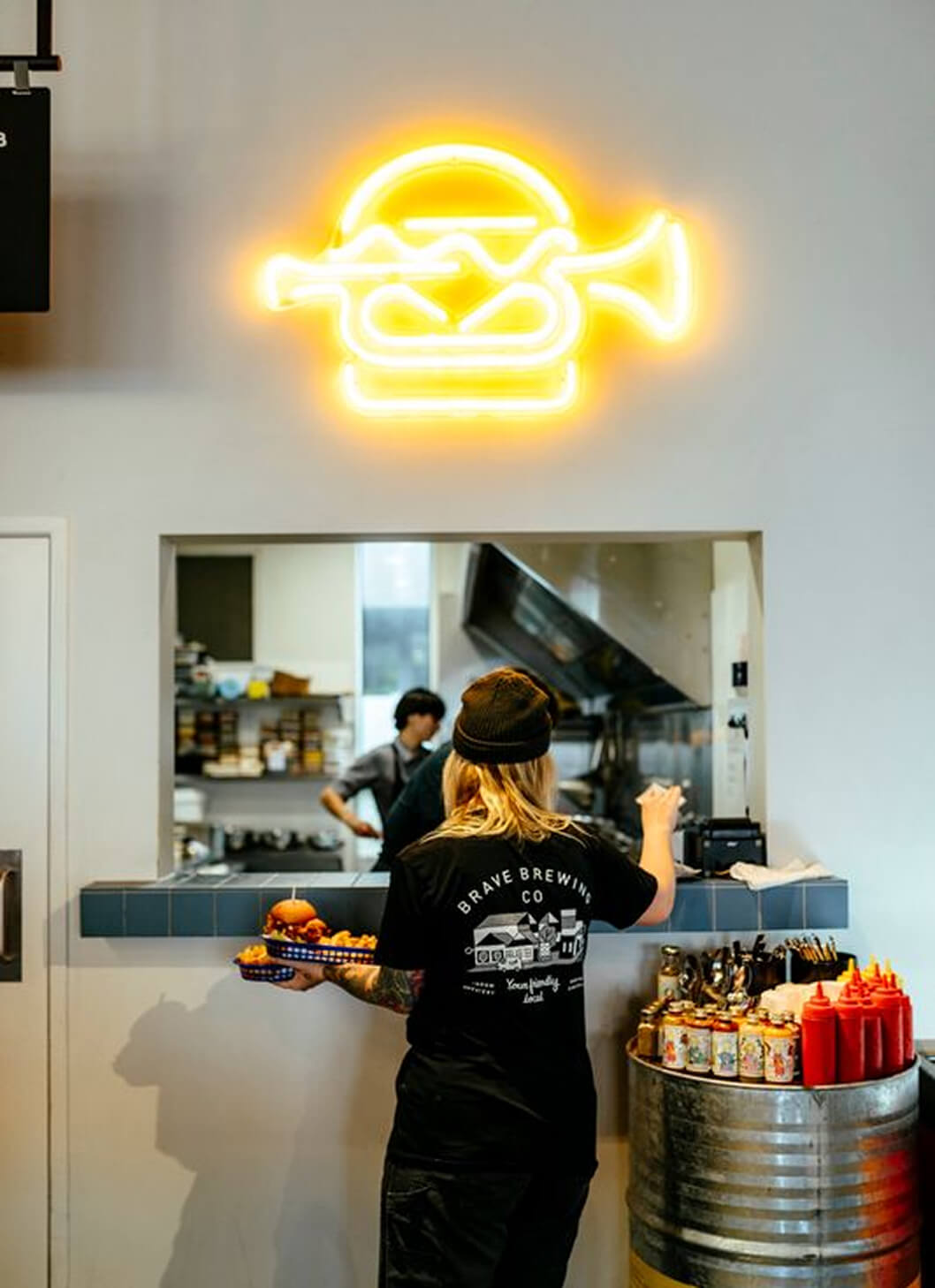Person holding a tray in the kitchen with Brave Brewing Co neon sign above them